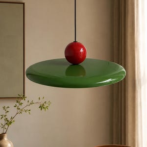 Könnte beinhalten: Eine Pendelleuchte mit einem glänzenden, olivgrünen, scheibenförmigen Lampenschirm, der an einem dünnen schwarzen Kabel hängt. Ein glattes, kugelförmiges rotes Dekorelement sitzt direkt auf der Mitte der grünen Scheibe und verbindet sie mit dem Kabel.