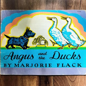 May include: A rectangular children's book titled Angus and the Ducks by Marjorie Flack. The cover features a black Scottish Terrier facing two white ducks, with a small green tree between them, set against a colorful rainbow and blue background.