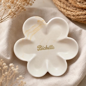 Peut inclure: Un vide-poche en forme de fleur de couleur blanc cassé avec de subtiles veines marbrées beige. Le centre du plat présente le mot Bichette imprimé dans une police cursive dorée. Le plat possède six pétales arrondis et une finition lisse.