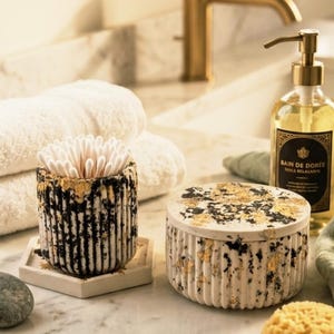 Puede incluir: Un recipiente cilíndrico lleno de bastoncillos de algodón blancos y un tarro a juego con tapa, ambos con un exterior blanco acanalado con motas negras y detalles de pan de oro. El recipiente se asienta sobre una base hexagonal blanca.