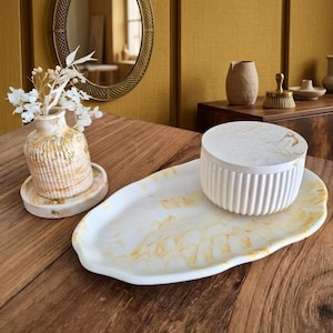 Peut inclure: Un plateau ovale en marbre blanc et doré aux bords irréguliers, accompagné d'un pot à couvercle assorti présentant des côtés cannelés verticaux. L'ensemble est présenté avec un petit vase contenant des fleurs séchées blanches sur un sous-verre.