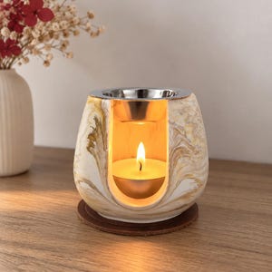Op de afbeelding: Een keramische oliebrander met een wit en goudkleurige marmeren afwerking, voorzien van een U-vormige opening met een brandend waxinelichtje. Bovenop bevindt zich een afneembaar zilverkleurig metalen schaaltje, rustend op een ronde bruine voet.