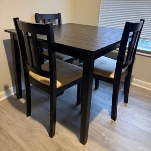 May include: A square dining table with a smooth black finish and four matching chairs. Each chair features a slatted back design and a seat cushion upholstered in a light beige fabric. The table has straight legs and a solid, flat tabletop surface.