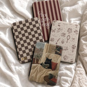 May include: Four e-reader cases with distinct designs: one with brown and white checkers and the letters EG, one with maroon and white vertical stripes and the text Hot girls read, one with cream and brown vintage icons, and one with a black cat reading.