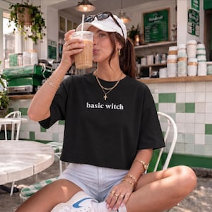 May include: A black short-sleeved t-shirt featuring the white text basic witch printed on the chest. The shirt has a loose, boxy fit and is paired with light-colored denim shorts, a white baseball cap, and layered gold-tone necklaces.