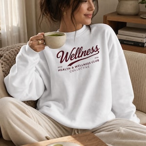 May include: A white crewneck sweatshirt featuring dark red text that reads Wellness ESTD 2026 HEALTH & WELLNESS CLUB COLLECTIVE. The design is printed on the chest of the long-sleeved garment, which has a ribbed collar, cuffs, and hemline.