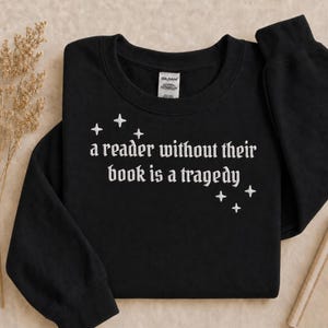 Pode incluir: Uma camisola preta de manga comprida com gola redonda, apresentando o texto bordado a branco "a reader without their book is a tragedy" e pequenos ícones de estrelas bordados. A peça inclui uma etiqueta de pescoço da marca Gildan.