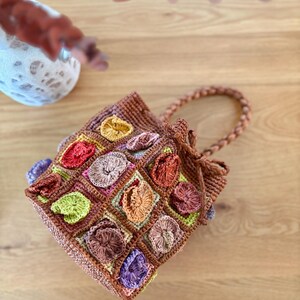 May include: A brown crochet bucket bag featuring a grid of square patches, each adorned with a raised, circular floral motif in various colors including red, yellow, green, purple, and orange. The bag has a braided handle and a drawstring closure.