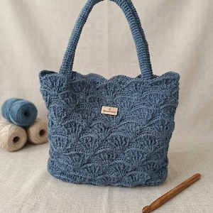 Könnte beinhalten: Eine staubblaue Häkeltasche mit einem sich wiederholenden Muschelmuster und zwei stabilen Henkeln. Ein kleines rechteckiges Holzschild ist mittig auf der Vorderseite angebracht, auf dem der Text "la handmade" in dunkler Schreibschrift eingraviert ist.