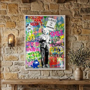 May include: A rectangular wall art print featuring a black and white illustration of Albert Einstein holding a sign that reads "LOVE IS THE ANSWER". The background is a vibrant, multi-colored graffiti collage with various text and cartoon motifs.