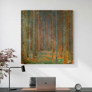 Op de afbeelding: Een vierkante wandkunstprint met een dicht bos met hoge, dunne boomstammen in bruine, oranje en groene tinten. Het kunstwerk hangt aan een witte muur boven een zwart bureau, waarop een laptop en een plant staan opgesteld.