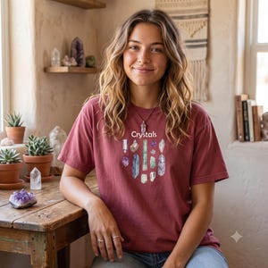 May include: A short-sleeved, crew-neck t-shirt in a muted brick red color. The front features a graphic illustration of various colorful crystals arranged in a cluster, with the word "Crystals" printed in white text above the crystal collection.
