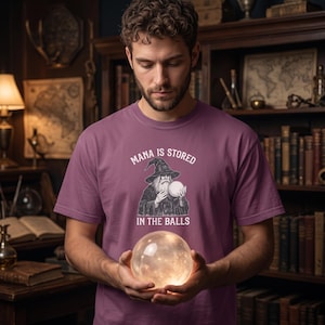 Può includere: Una t-shirt a maniche corte color malva con una stampa grafica di un mago che tiene in mano un globo luminoso. Il testo sopra il mago recita "MANA IS STORED" e il testo sottostante "IN THE BALLS" in lettere maiuscole bianche.
