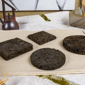 May include: A set of four dark grey, porous coasters with a rough, volcanic-like texture. The collection includes two square coasters and two circular coasters, all featuring a flat surface and a consistent, dark, earthy, and pitted appearance.