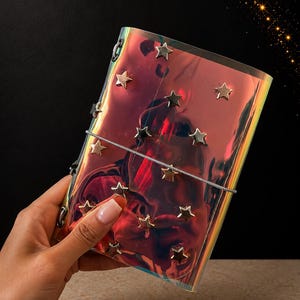 May include: A rectangular notebook with a reflective, iridescent, holographic cover featuring a series of small, metallic, five-pointed star studs scattered across the front. A thin, light-colored elastic band wraps horizontally around the center.