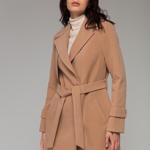 May include: A camel-colored wool-blend coat featuring a wide notched lapel, long sleeves with cuff straps, and a matching tie belt at the waist. The coat is worn over a cream-colored ribbed turtleneck sweater, creating a layered look.