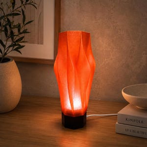 May include: A tall, orange, translucent lamp with a textured, folded, geometric surface and a hexagonal base. The lamp emits a warm, glowing light from within, casting a soft illumination upward through its unique, sculptural, vertical structure.