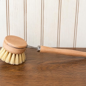 May include: A dish brush with a round wooden head, light-colored natural bristles, and a long wooden handle. The brush features a metal connector between the head and the handle, and a small metal loop at the end of the handle for hanging.