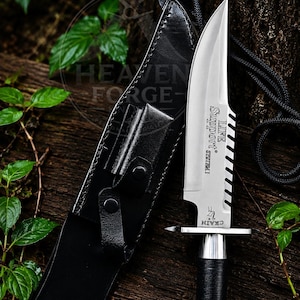 May include: A silver-toned survival knife with a black wrapped handle and a serrated spine. The blade is engraved with the text 'LIFE SUPPORT SYSTEM I' and 'CRAIN'. It is accompanied by a black leather sheath with a snap closure and a black cord.