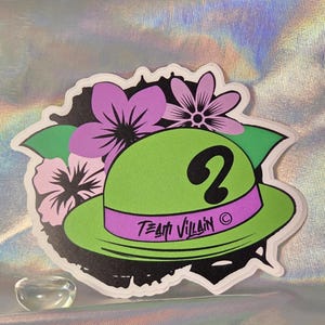 May include: A sticker featuring a green hat with a purple band and a black question mark. The hat is surrounded by purple and pink flowers and the words "Team Villain" are printed on the band.