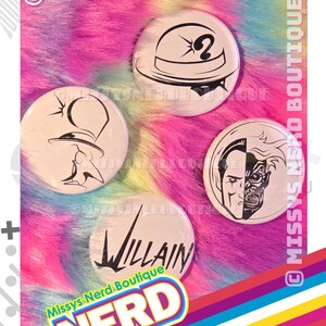 May include: Four circular pin-back buttons featuring black line art designs on a white background. One button shows a bowler hat with a question mark, one shows a stylized face, one shows a split-face character, and one features the word Villain.