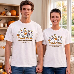 May include: A white short-sleeve t-shirt featuring a graphic of three ghosts dressed as ducks holding books and a pumpkin bucket. The text reads CURRENTLY HAUNTING THE LIBRARY and Too Ghoul for Fiction in brown lettering below the illustration.