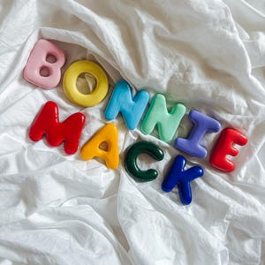 May include: A collection of individual, glossy, three-dimensional letters spelling out the words BONNIE MACK. The letters are crafted in various vibrant colours including pink, yellow, red, orange, light blue, mint green, purple, dark green, and blue.
