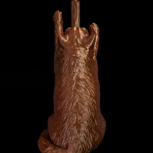 May include: A brown, textured figurine of a cat sitting upright with its front paws raised high. The cat is positioned behind a tall, brown, cylindrical post that features a small, yellow, bird-like figure perched at the very top of the structure.