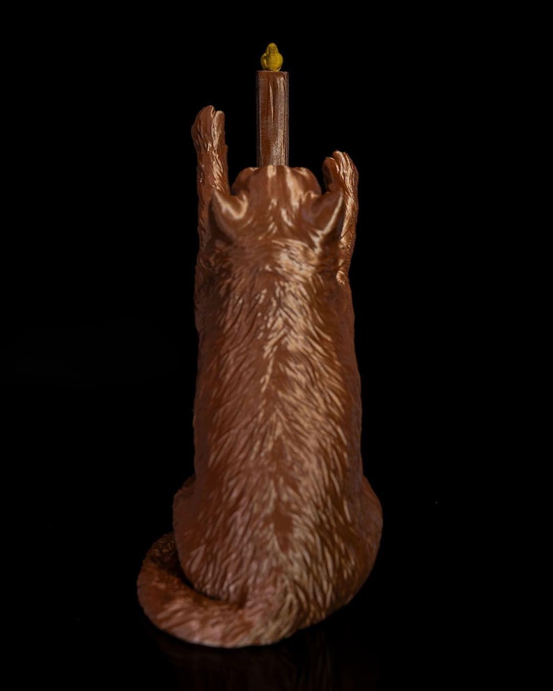 May include: A brown, textured figurine of a cat sitting upright with its front paws raised high. The cat is positioned behind a tall, brown, cylindrical post that features a small, yellow, bird-like figure perched at the very top of the structure.