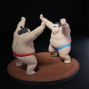 May include: A figurine set featuring two sumo wrestlers in a grappling stance on a circular brown base. One wrestler wears a red mawashi, while the other wears a blue mawashi. Both figures have dark hair styled in a topknot and are posed with arms raised.