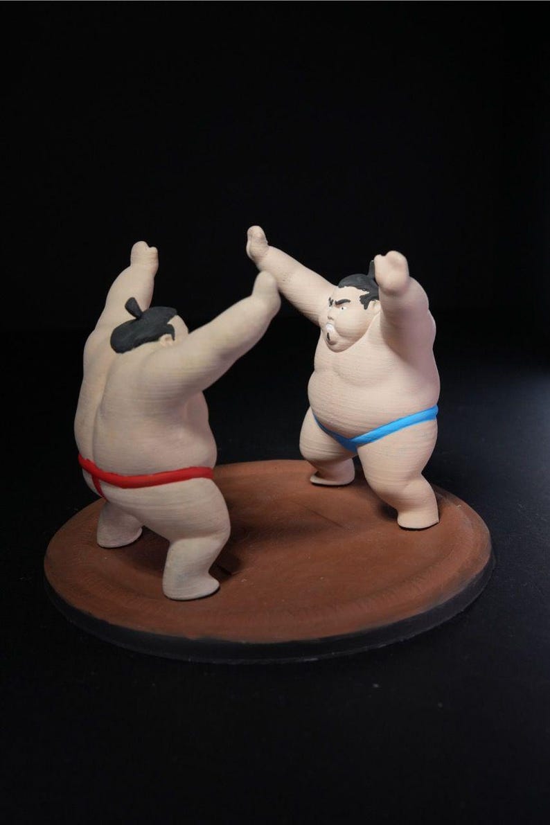 May include: A figurine set featuring two sumo wrestlers in a grappling stance on a circular brown base. One wrestler wears a red mawashi, while the other wears a blue mawashi. Both figures have dark hair styled in a topknot and are posed with arms raised.