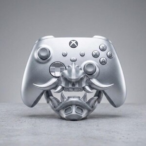 Puede incluir: Un mando inal&aacute;mbrico Xbox de color plata met&aacute;lico con una m&aacute;scara de samur&aacute;i tridimensional personalizada. La m&aacute;scara cubre la parte inferior del mando y muestra colmillos esculpidos, un protector nasal y una pieza para la barbilla.