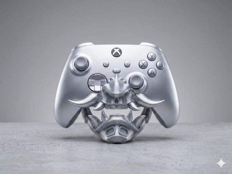 Puede incluir: Un mando inal&aacute;mbrico Xbox de color plata met&aacute;lico con una m&aacute;scara de samur&aacute;i tridimensional personalizada. La m&aacute;scara cubre la parte inferior del mando y muestra colmillos esculpidos, un protector nasal y una pieza para la barbilla.