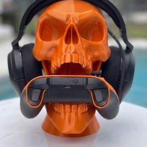 May include: A bright orange 3D-printed human skull model serving as a stand. The skull is wearing a pair of black over-ear gaming headphones and has a black and orange video game controller positioned horizontally within its open jaw.