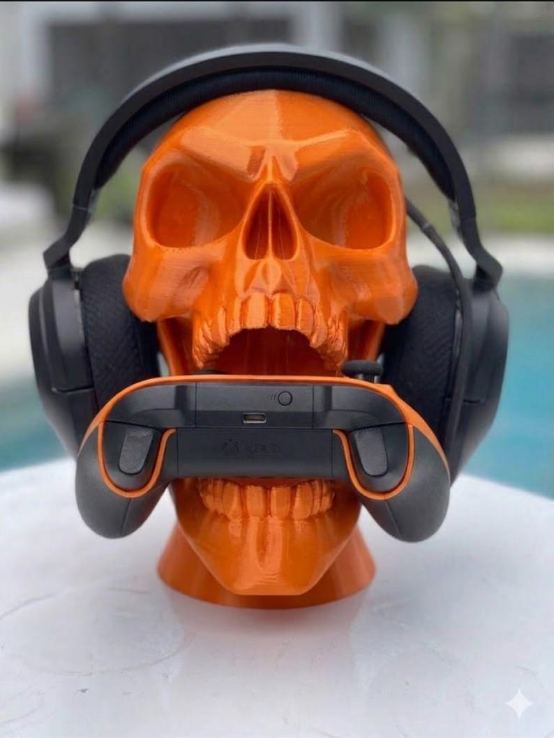 May include: A bright orange 3D-printed human skull model serving as a stand. The skull is wearing a pair of black over-ear gaming headphones and has a black and orange video game controller positioned horizontally within its open jaw.