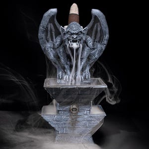 Puede incluir: Un quemador de incienso de g&aacute;rgola color piedra gris con una criatura alada posada sobre un pedestal escalonado. Un cono de incienso marr&oacute;n se sit&uacute;a detr&aacute;s de la g&aacute;rgola, liberando humo blanco que fluye desde la boca hacia la base.