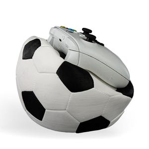 Op de afbeelding: Een witte videogamecontroller die in een zwart-witte voetbalvormige houder rust. De controller is voorzien van zwarte analoge sticks, een richtingspad en gekleurde actieknoppen. De voetbal heeft een getextureerd oppervlak met stiksels.