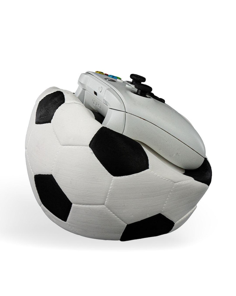 Op de afbeelding: Een witte videogamecontroller die in een zwart-witte voetbalvormige houder rust. De controller is voorzien van zwarte analoge sticks, een richtingspad en gekleurde actieknoppen. De voetbal heeft een getextureerd oppervlak met stiksels.