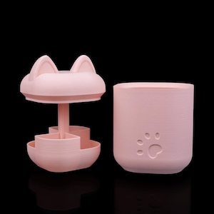 May include: A light pink plastic storage container with a cat-ear lid and a central post featuring a multi-compartment rotating tray. The cylindrical base of the container features a debossed paw print design on the exterior surface.