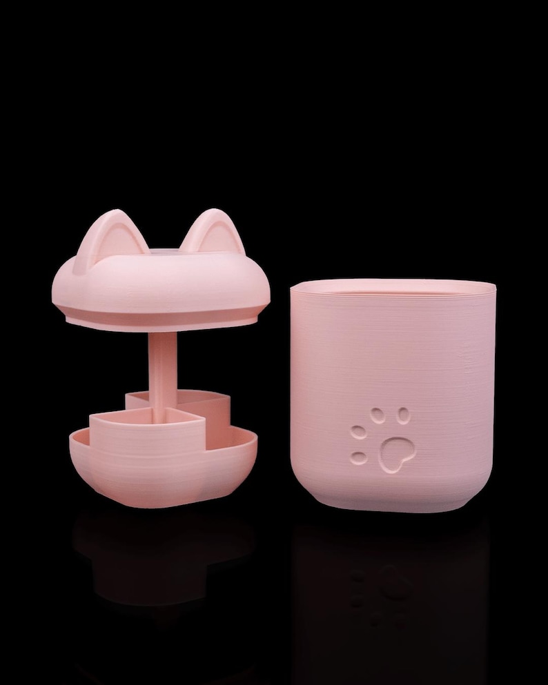 May include: A light pink plastic storage container with a cat-ear lid and a central post featuring a multi-compartment rotating tray. The cylindrical base of the container features a debossed paw print design on the exterior surface.