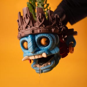 May include: A blue, mask-like planter featuring a brown, crown-shaped top with rope-like detailing. The face has large, glowing orange eyes, prominent white fangs, and a dark mouth with a clear liquid dripping from it. A green cactus is visible inside.