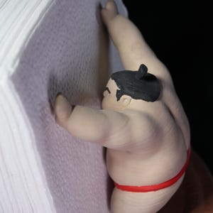 May include: A figurine of a sumo wrestler with black hair tied in a topknot, wearing a red mawashi, pushing against a stack of white paper sheets. The wrestler has a light skin tone and is positioned in a forward-leaning stance against the paper.