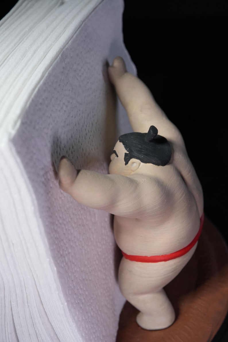 May include: A figurine of a sumo wrestler with black hair tied in a topknot, wearing a red mawashi, pushing against a stack of white paper sheets. The wrestler has a light skin tone and is positioned in a forward-leaning stance against the paper.
