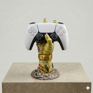 May include: A white and black video game controller resting on a yellow, textured hand-shaped stand. The hand features a gold-colored wristband, visible stitches, and a small insect detail, emerging from a rocky, earth-toned circular base.