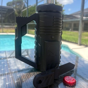 Pode incluir: Uma pega de garrafa preta impressa em 3D com um design t&aacute;tico, apresentando um emblema de caveira moldado e um padr&atilde;o de bandeira americana. A pega est&aacute; fixada ao gargalo de uma garrafa de vidro transparente, com uma ferramenta preta e uma tampa Smirnoff.