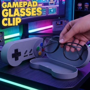 May include: A gray, controller-shaped glasses case with a black directional pad and colored buttons, featuring the text SELECT and START. The case is open, revealing a pair of black-rimmed glasses being held above the interior compartment.