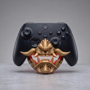 Puede incluir: Un mando de videojuegos inal&aacute;mbrico negro con un accesorio de m&aacute;scara de samur&aacute;i impreso en 3D de color dorado. La m&aacute;scara cubre la parte inferior del mando y muestra colmillos prominentes y un acabado met&aacute;lico detallado y texturizado.