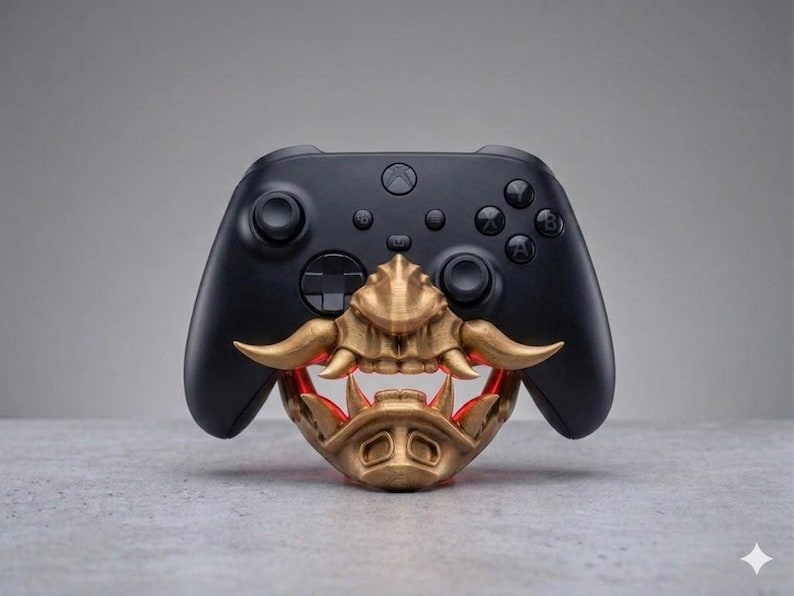 Puede incluir: Un mando de videojuegos inal&aacute;mbrico negro con un accesorio de m&aacute;scara de samur&aacute;i impreso en 3D de color dorado. La m&aacute;scara cubre la parte inferior del mando y muestra colmillos prominentes y un acabado met&aacute;lico detallado y texturizado.
