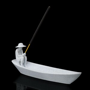 May include: A light gray textured incense holder shaped like a small boat with a seated figure wearing a wide-brimmed hat. The figure holds a thin yellow rod that supports a long black incense stick angled upward against a solid black background.