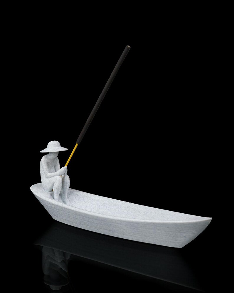 May include: A light gray textured incense holder shaped like a small boat with a seated figure wearing a wide-brimmed hat. The figure holds a thin yellow rod that supports a long black incense stick angled upward against a solid black background.
